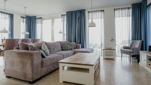 Een ruime woonkamer met een grote bank, een salontafel en een fauteuil. Er zijn grote ramen met blauwe gordijnen, een lamp en een kleine tafel.