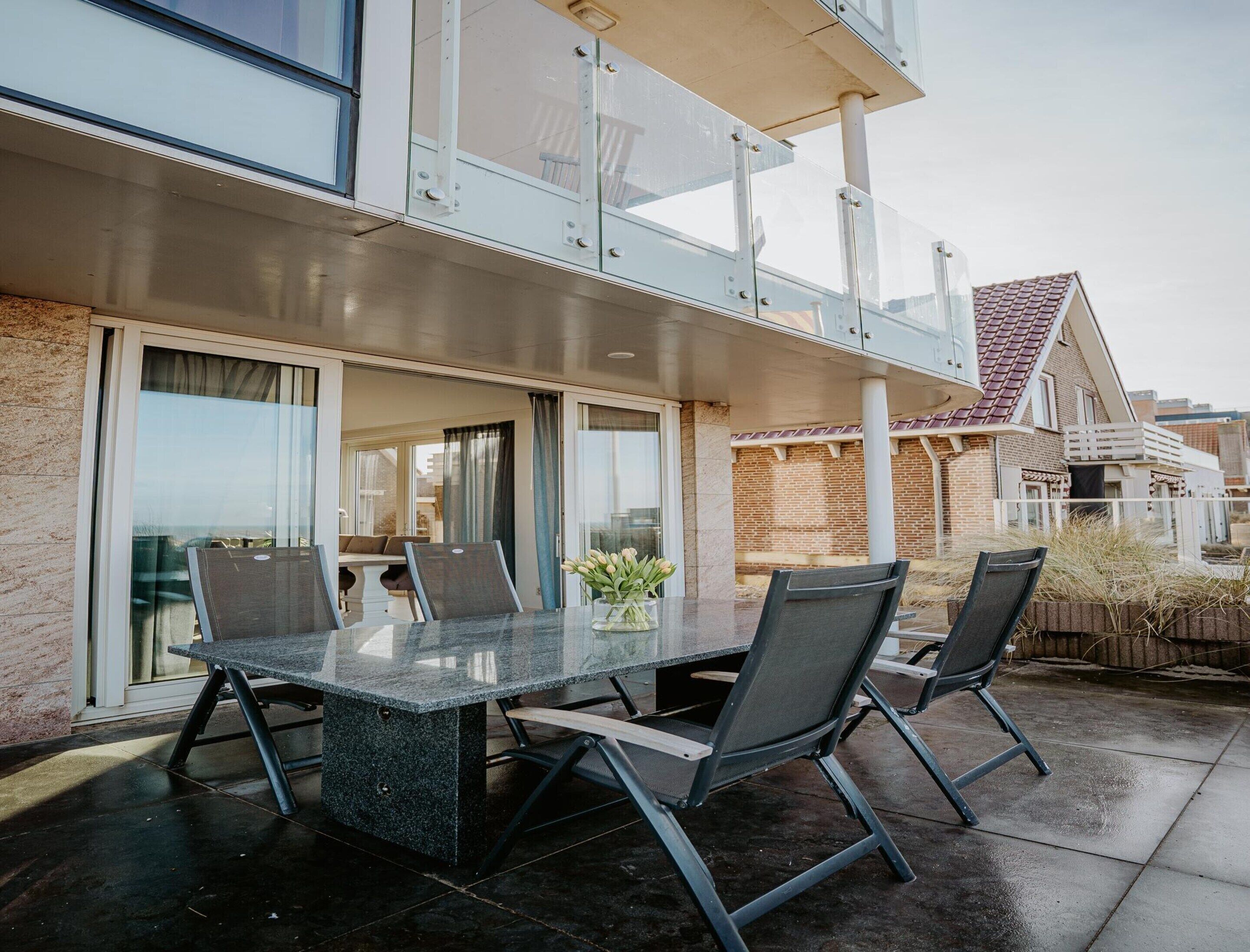 An outdoor patio with a glass-top table and four chairs, a potted plant, and a view of a residential area.