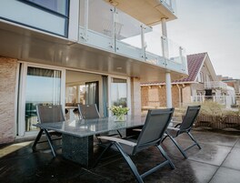 An outdoor patio with a glass-top table and four chairs, a potted plant, and a view of a residential area.