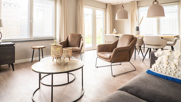 A modern living room with a brown leather sofa, a round coffee table, and a dining area with a wooden table and chairs. - Spacious Bungalow in the Cosy Village (De Cocksdorp)