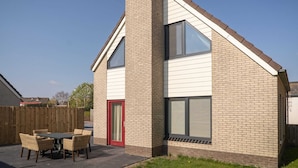 A modern house with a brick exterior, a red door, and a patio with wicker furniture. - Spacious Bungalow in the Cosy Village (De Cocksdorp)