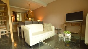 A modern living room with a white sofa, a glass coffee table, a TV on a wooden stand, and a kitchen area in the background.