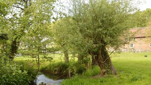 A rural landscape with a stream, trees, and a building in the background.