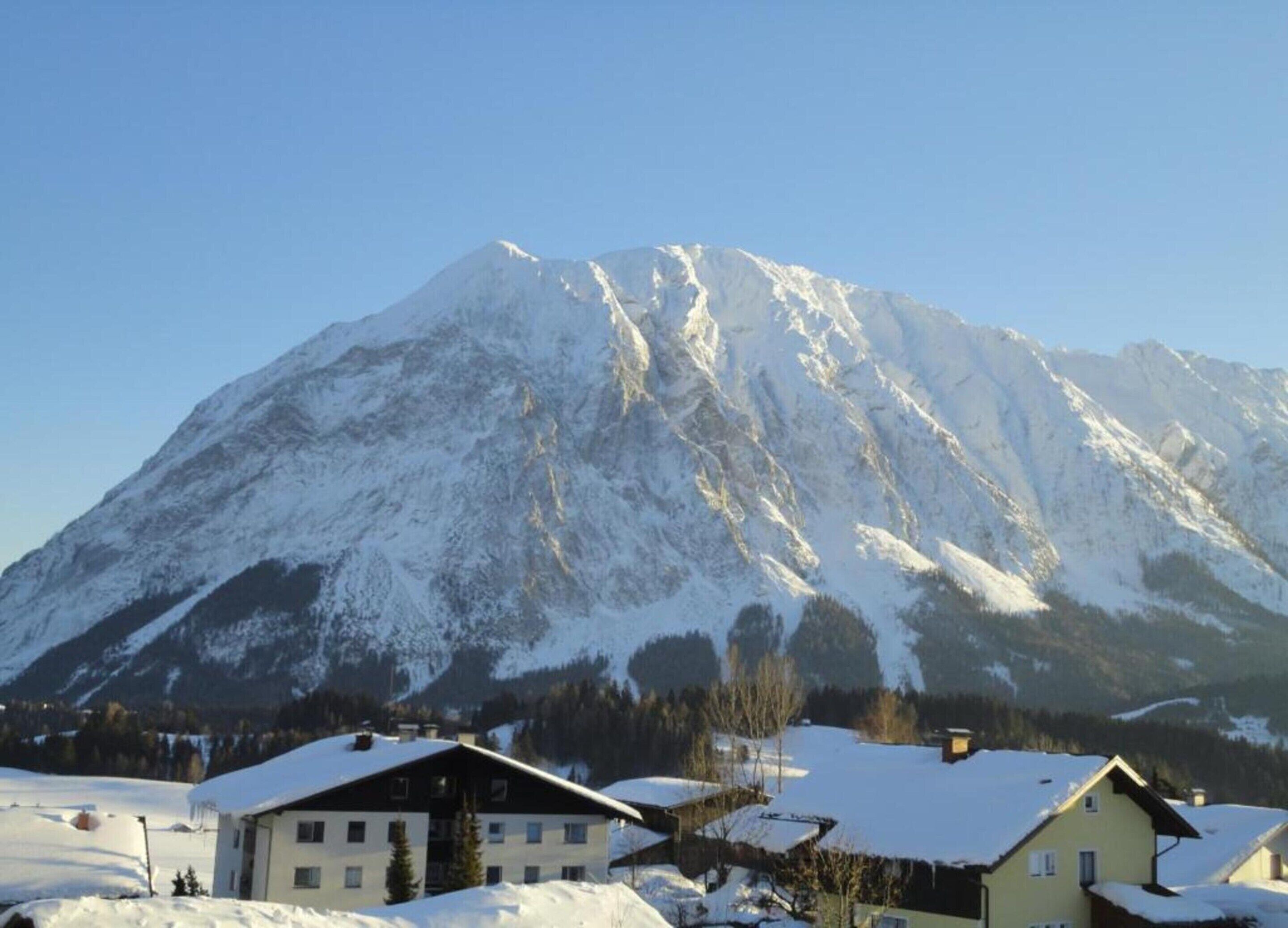 A snow-covered mountain with a clear sky. There are several buildings with snow on their roofs in the foreground.