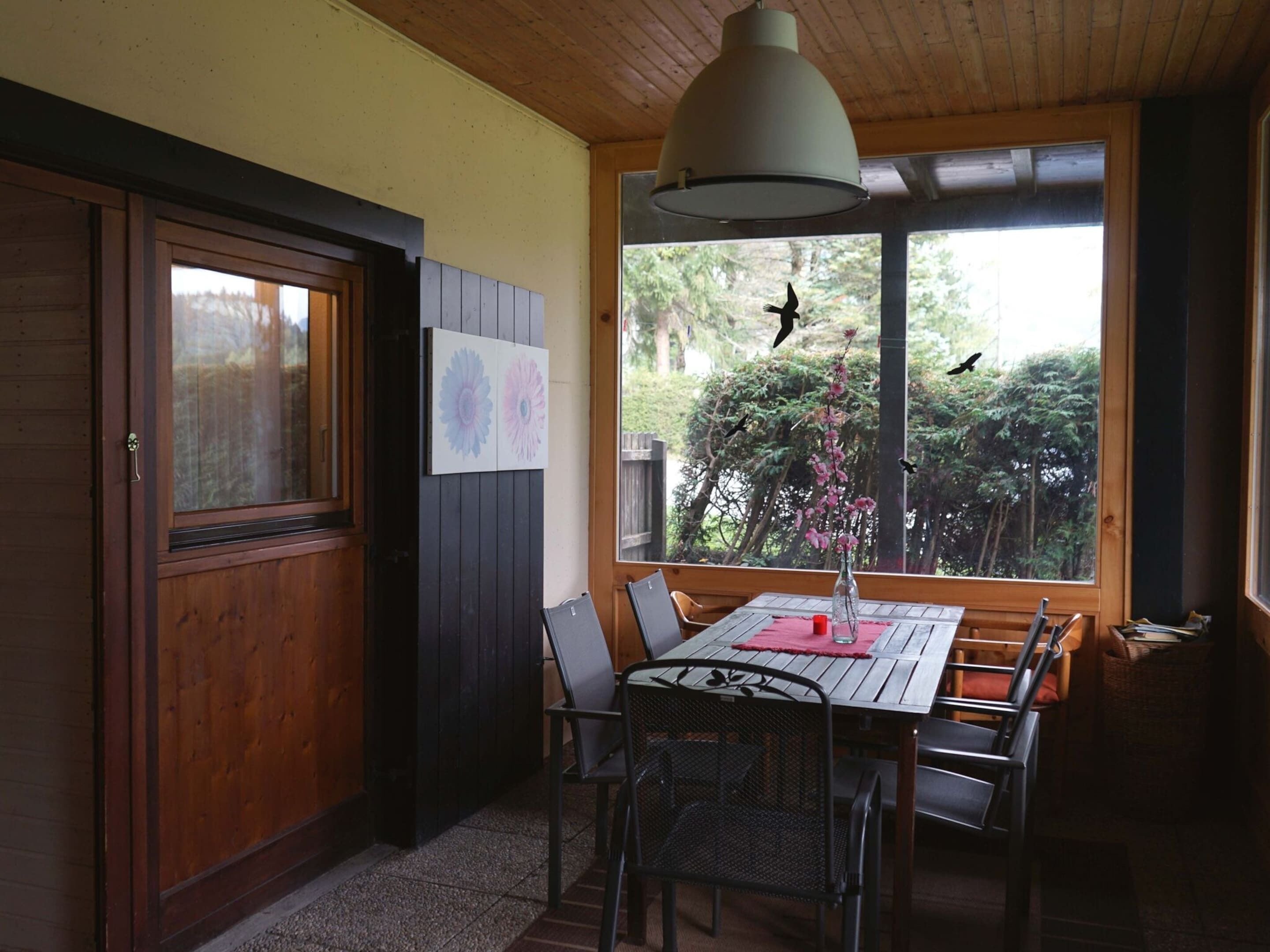 Un espace repas avec une table et des chaises en bois, un luminaire suspendu et une vue sur les arbres et les oiseaux, visible à travers de grandes fenêtres.