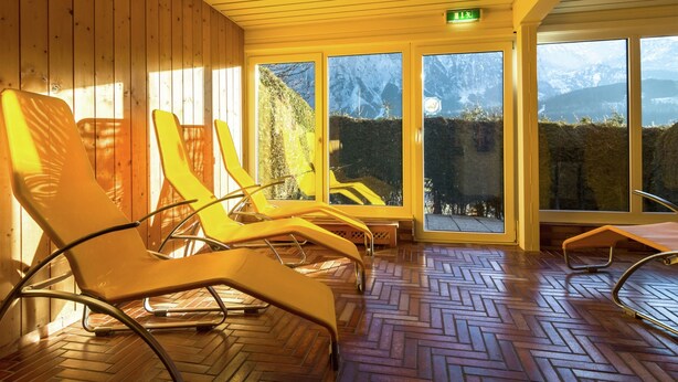 A room with wooden walls and a herringbone floor, featuring several yellow lounge chairs and large windows offering a mountain view. - Apartment in Tauplitz Near Ski Lift (Bad Mitterndorf)
