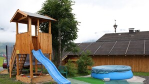 A wooden play structure with a slide and a small tower, surrounded by a grassy area and a building with a sloped roof. - Cozy Apartment in Embach Austria near Ski Area (Lend)