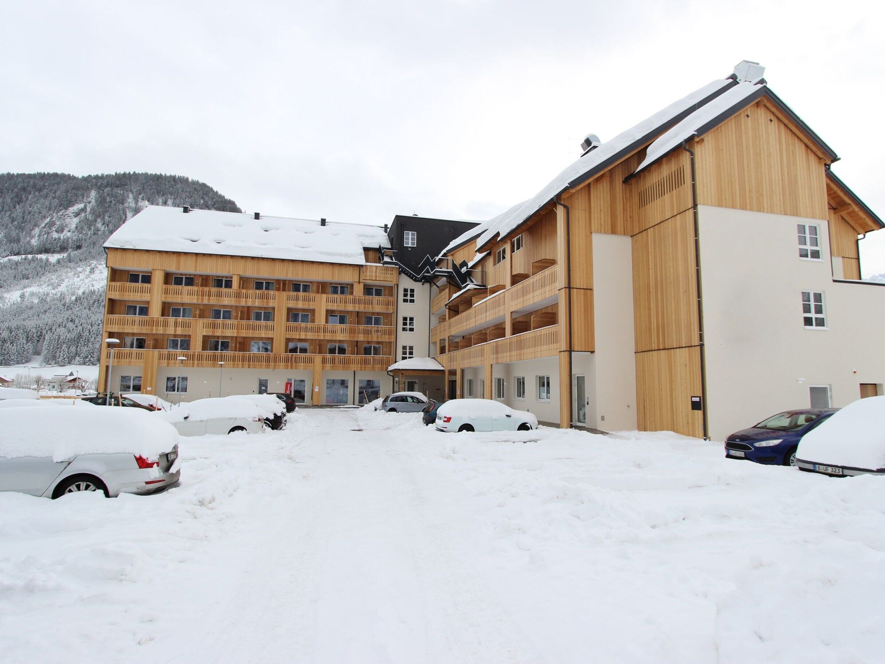 A multi-story wooden building with balconies, surrounded by snow-covered cars and a mountainous backdrop.