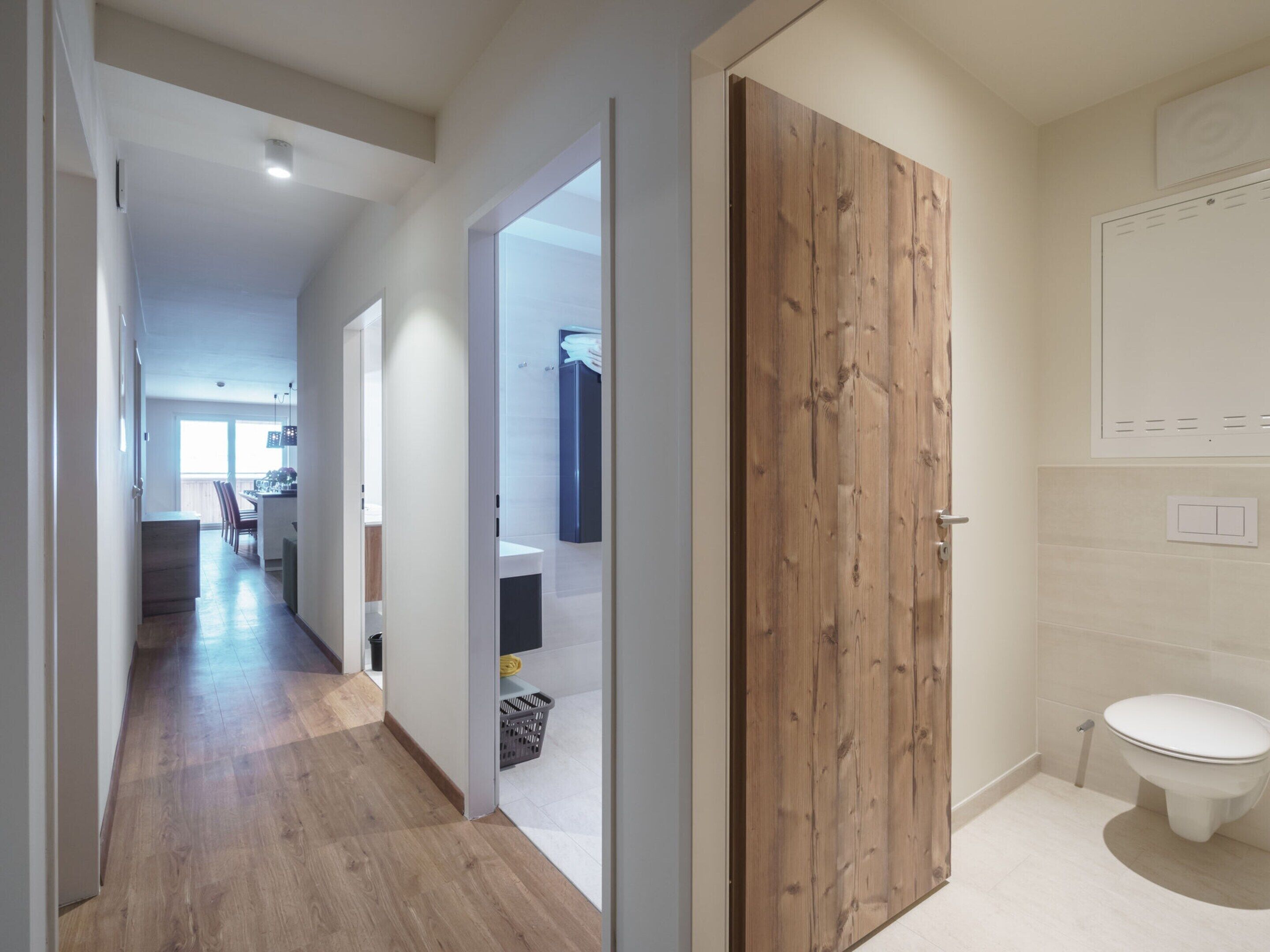 A modern apartment with a wooden door, a bathroom with a toilet, and a kitchen area visible through an open door.