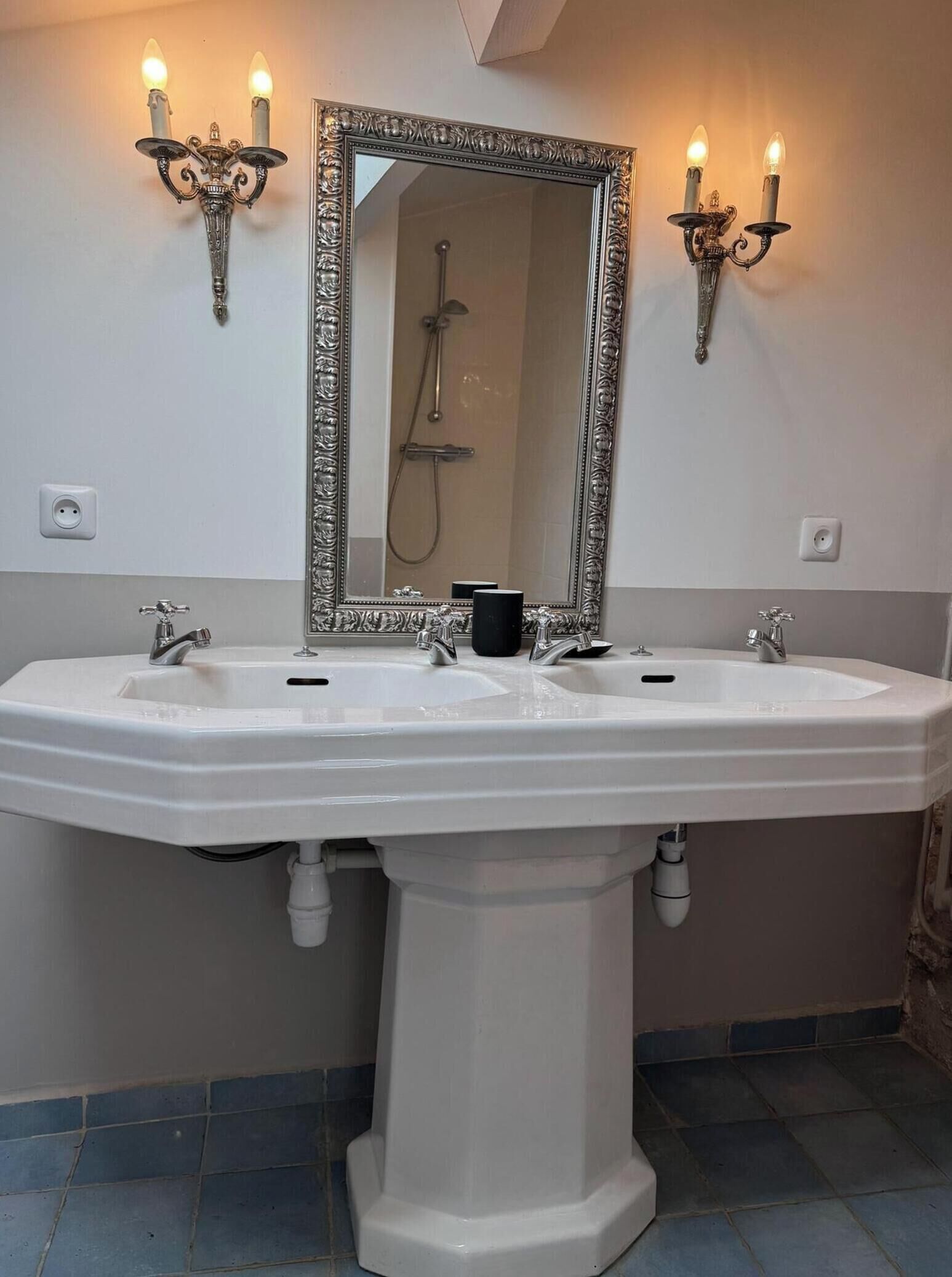 A bathroom with a large white pedestal sink, two faucets, a mirror with an ornate frame, and wall-mounted sconces.