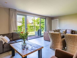 A modern living room with a sofa, a wicker chair, a wooden coffee table, and a view of a garden through large sliding glass doors. - Detached House Zeeland Near Beach (Kamperland)