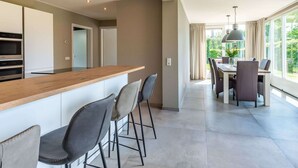A modern kitchen with a wooden countertop and bar stools, a dining area with a table and chairs, and large windows offering a view of greenery. - Detached House Zeeland Near Beach (Kamperland)