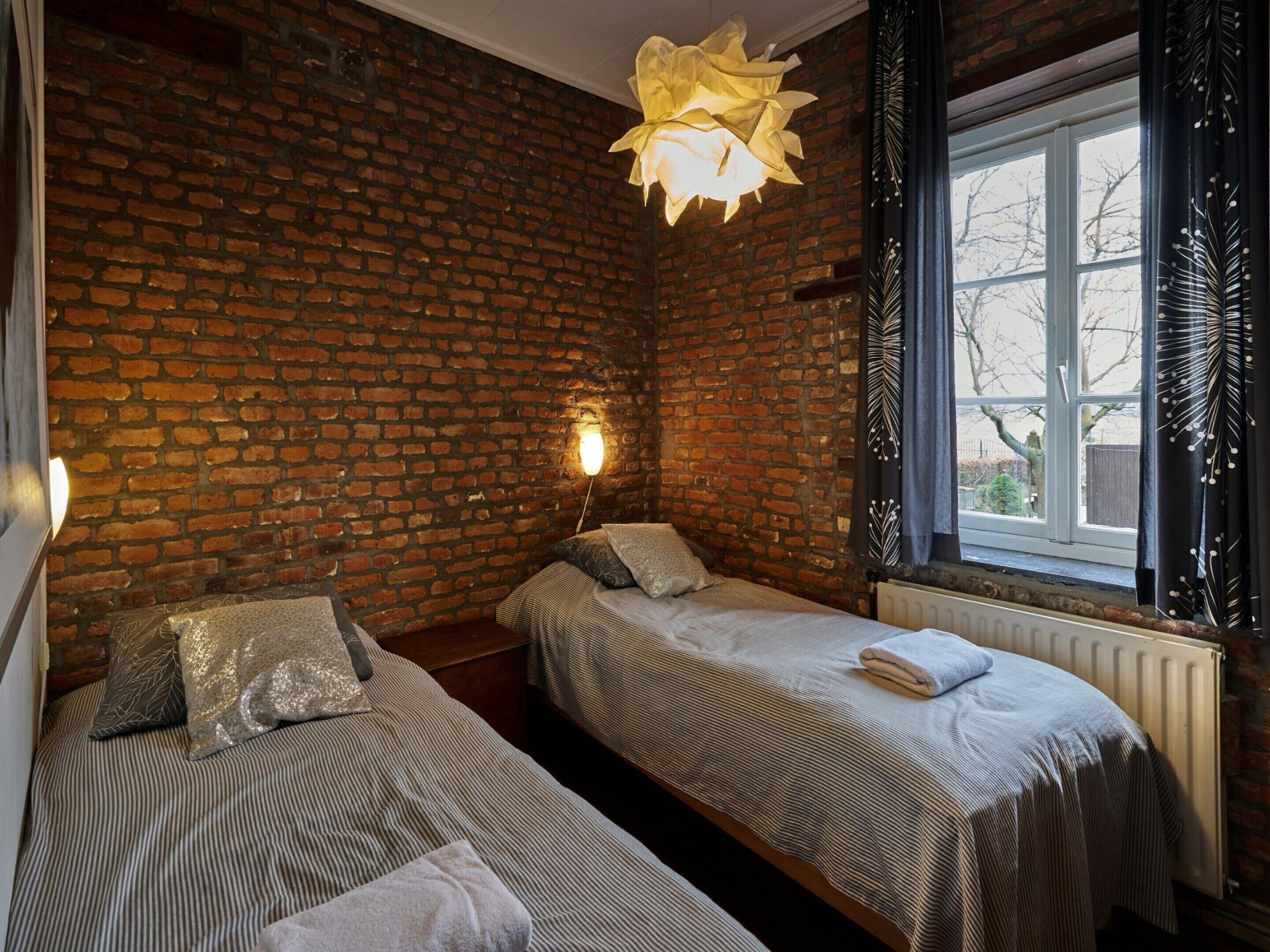 A room with a brick wall, two beds with striped bedding, a hanging light fixture, and a window with curtains.