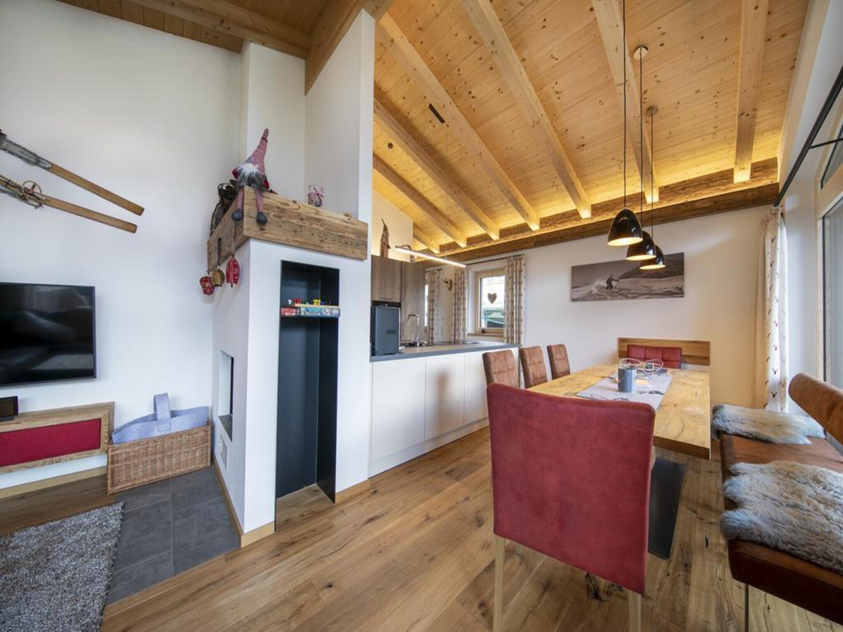 A modern interior with a wooden ceiling, a dining area with red chairs, a kitchen with a built-in fridge, and a TV mounted on the wall.