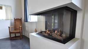 A room with a glass fireplace, a wooden chair, and a window with curtains. - Seaside Villa With Garden (Tzummarum)