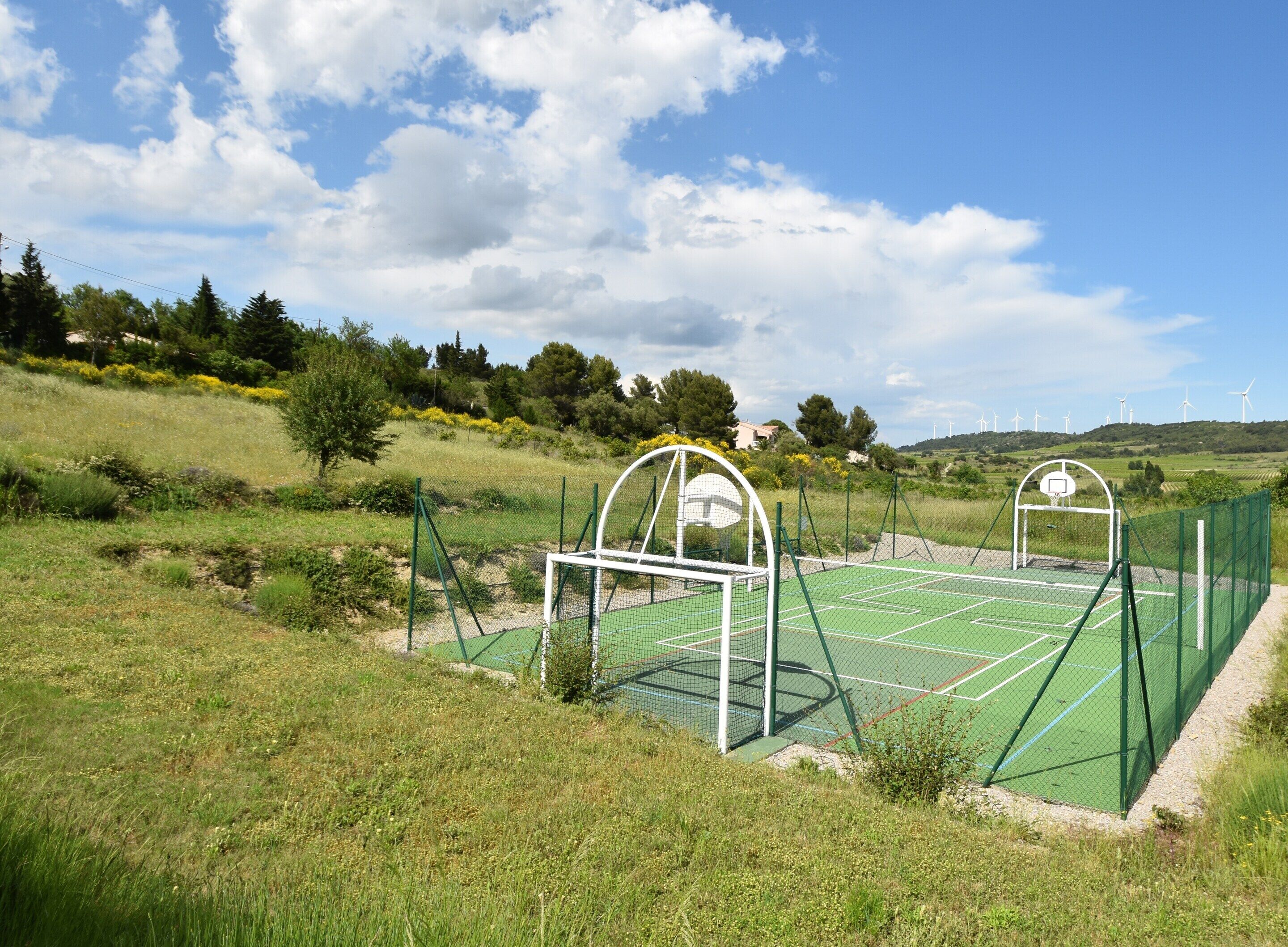 An outdoor tennis court with a green surface, surrounded by a green fence, and wind turbines in the background.