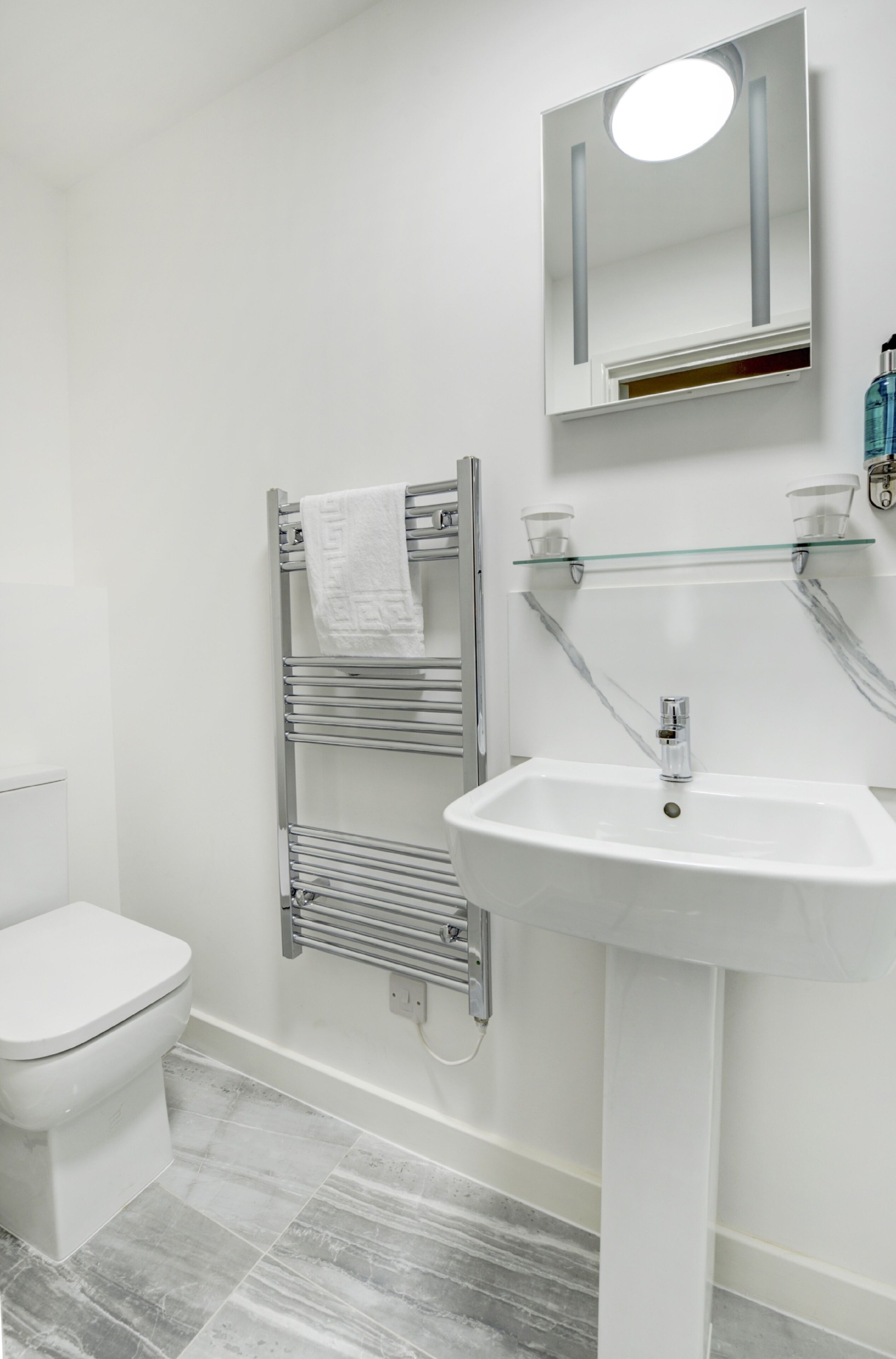 Een moderne badkamer met een wit toilet, een chroom handdoekrek, een glazen plank en een witte wastafel.