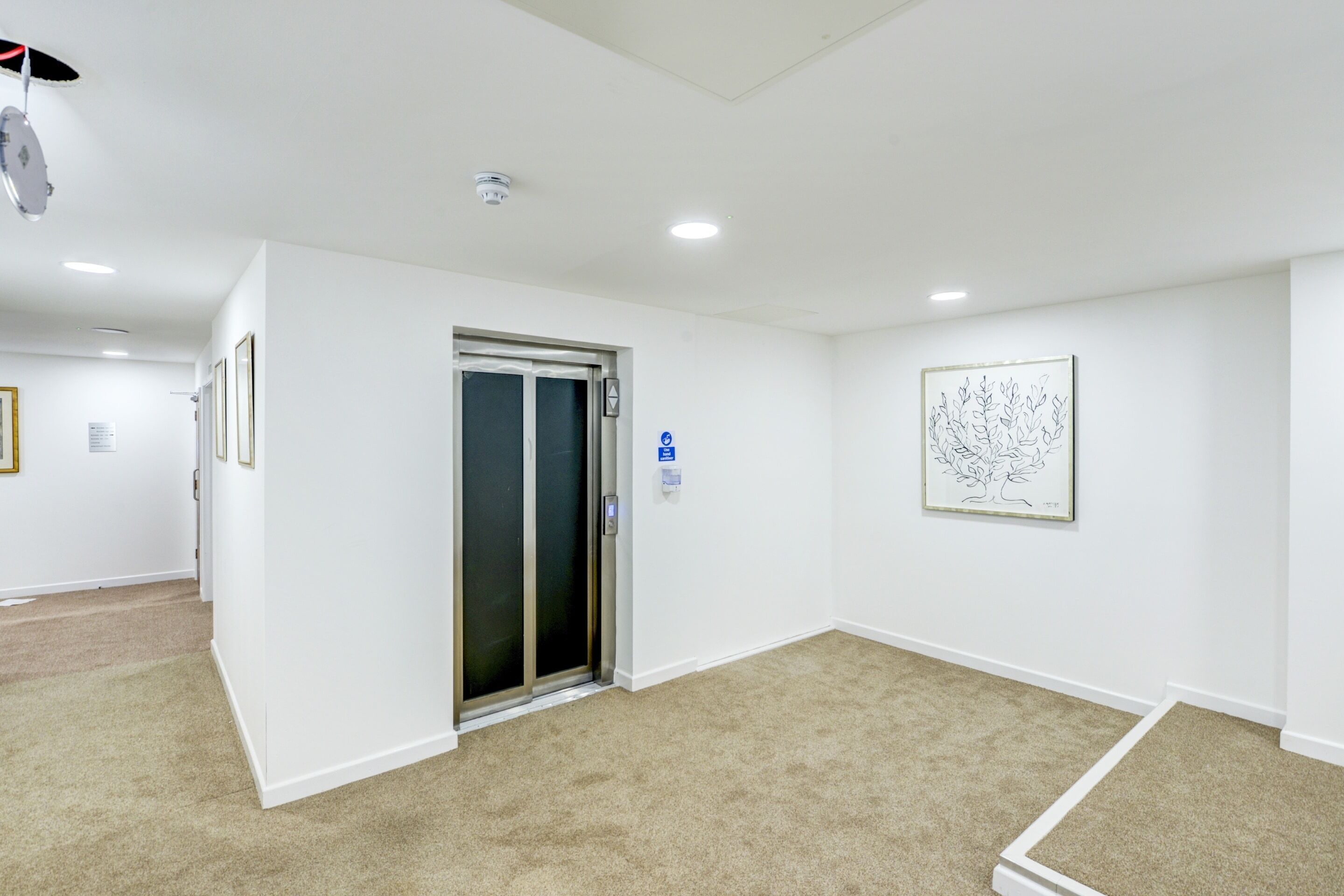Un couloir propre et moderne, doté d’un sol recouvert de moquette, d’une porte avec un panneau de verre et d’une œuvre d’art encadrée accrochée au mur.