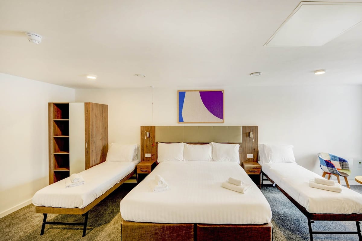 Une chambre d’hôtel avec trois lits simples, une armoire en bois, un grand tableau abstrait et une chaise avec un coussin coloré.