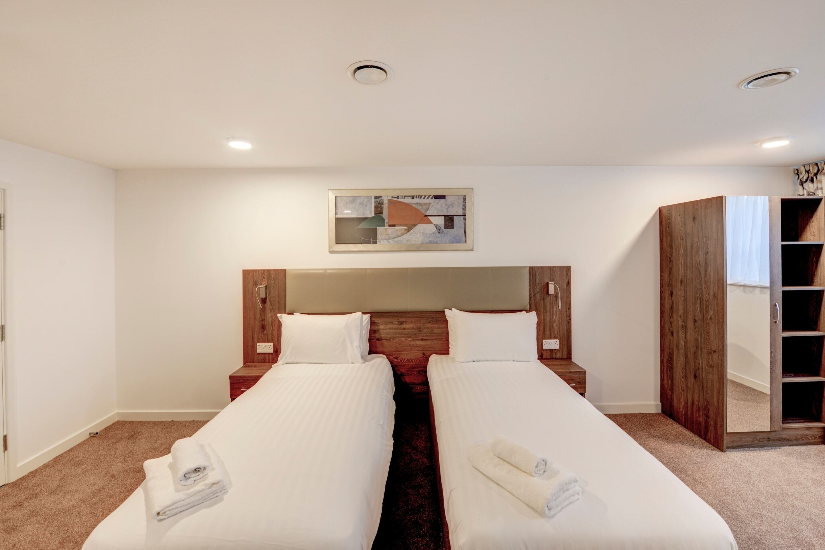 A hotel room with two single beds, a wooden headboard, a large mirror, and a framed abstract artwork on the wall.