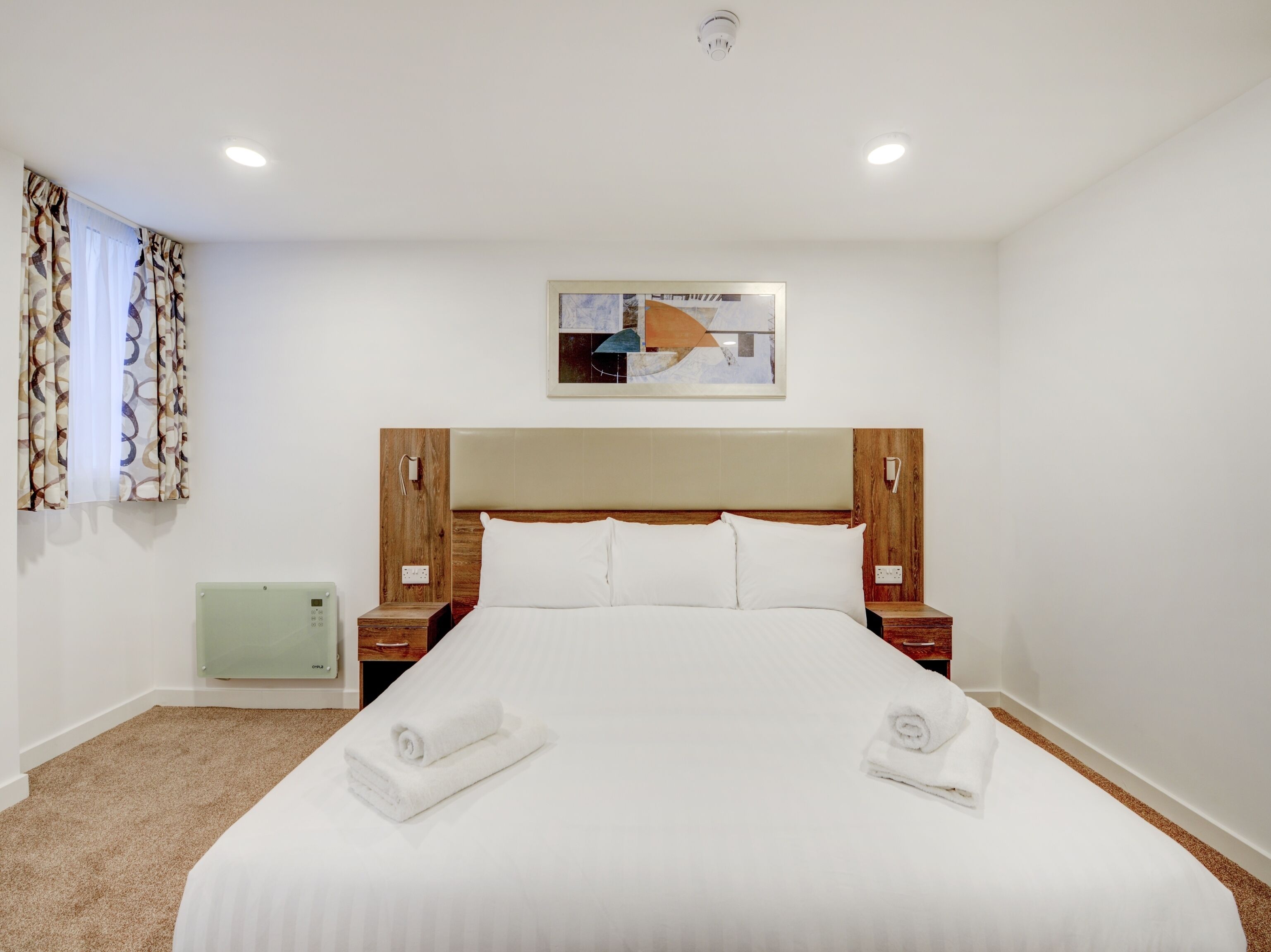 Habitación de hotel con una cama grande, cabecera de madera, dos mesitas de noche, un calentador verde y una ventana con cortinas estampadas.