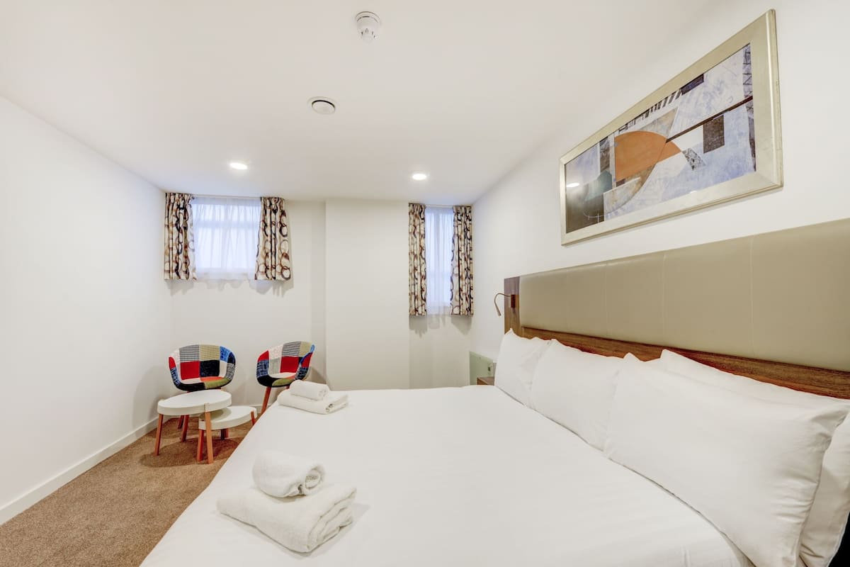 Une chambre d’hôtel avec un grand lit, deux chaises, un tableau au mur et une fenêtre avec des rideaux.