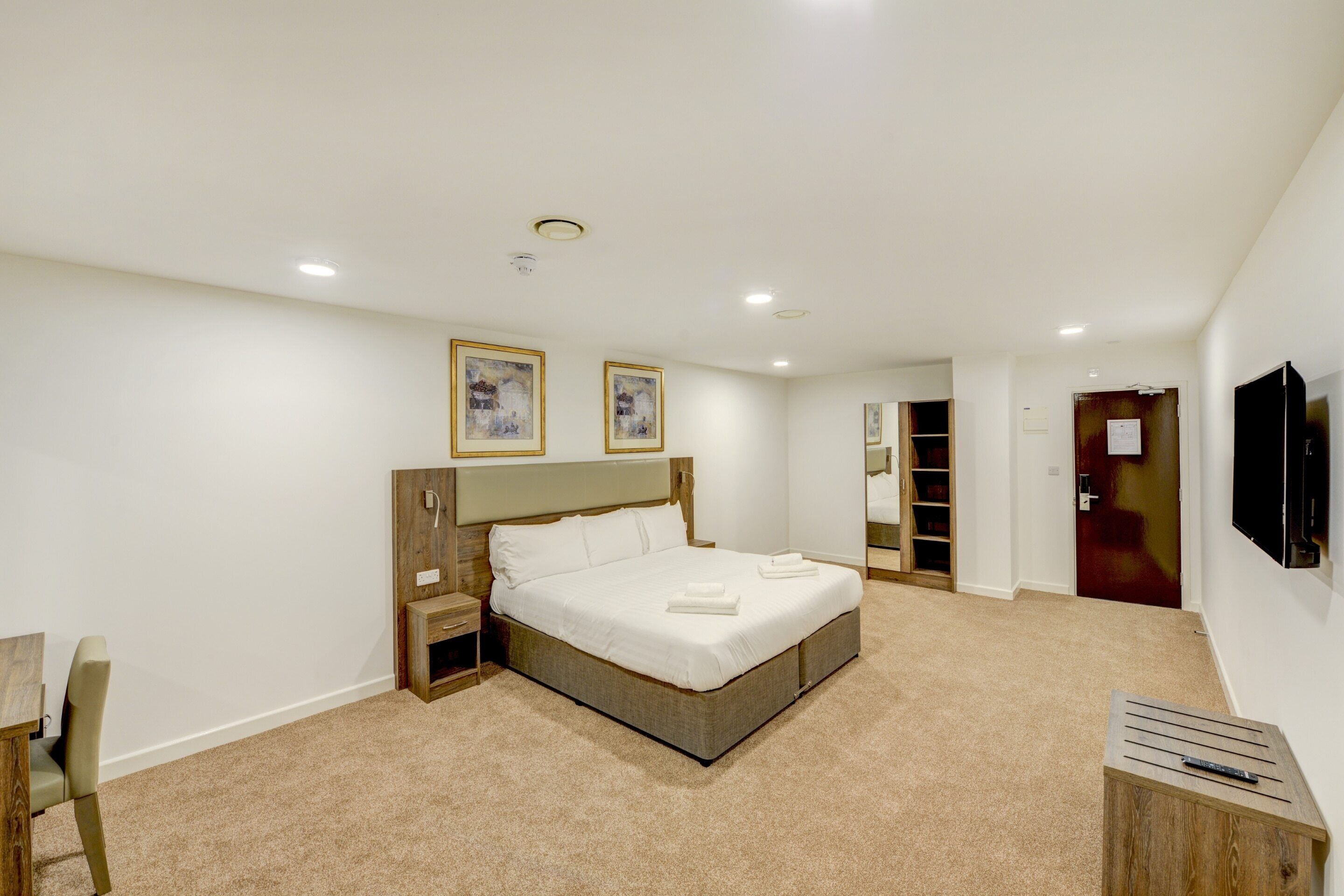 A bedroom with a large bed, a wooden nightstand, a bedside table, a chair, a TV mounted on the wall, and a door leading to another room.