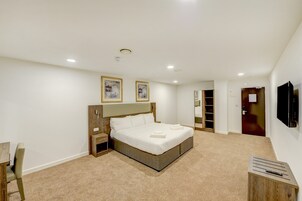A bedroom with a large bed, a wooden nightstand, a bedside table, a chair, a TV mounted on the wall, and a door leading to another room.