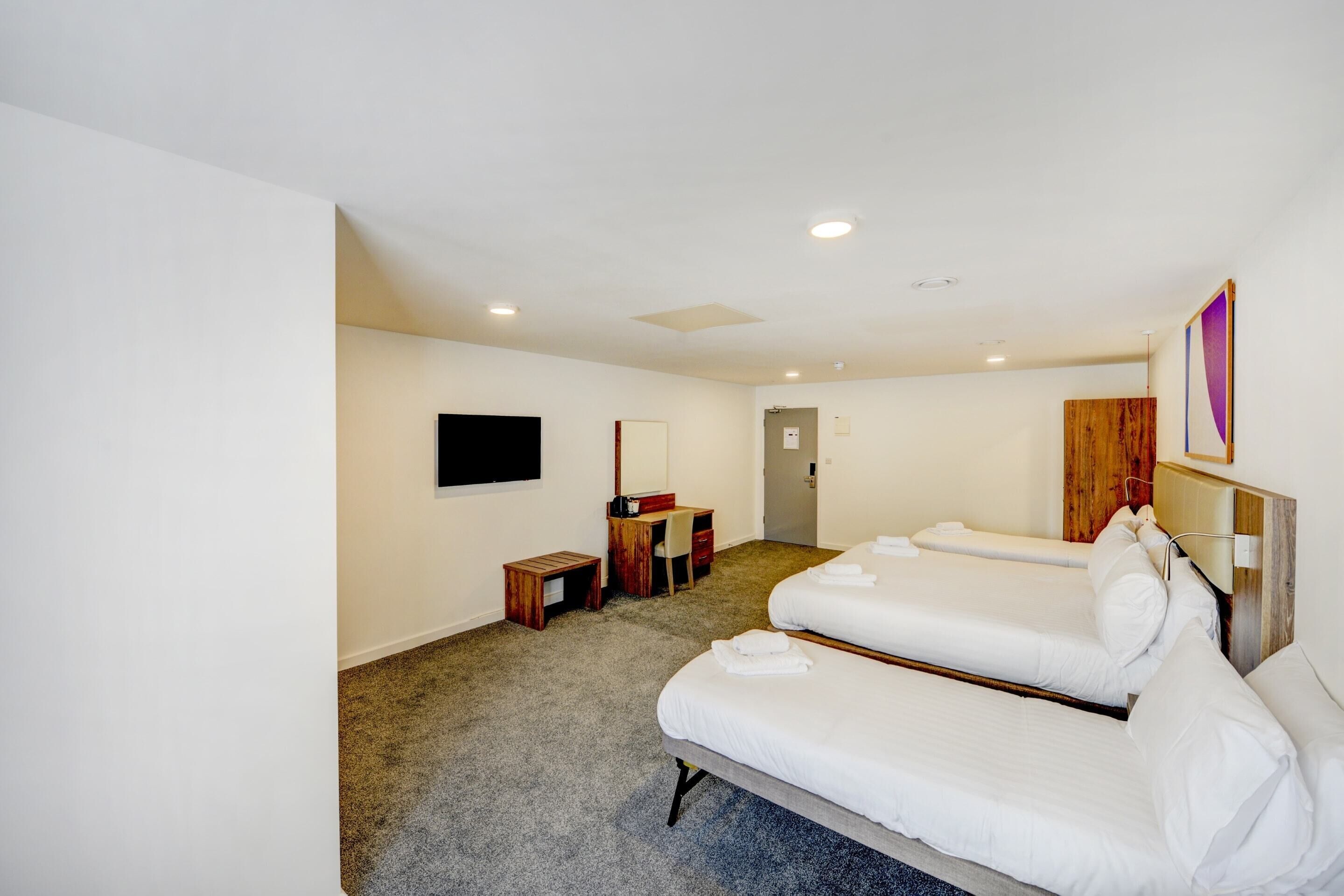 Quarto de hotel com duas camas, uma escrivaninha, uma cadeira, uma TV e uma porta.
