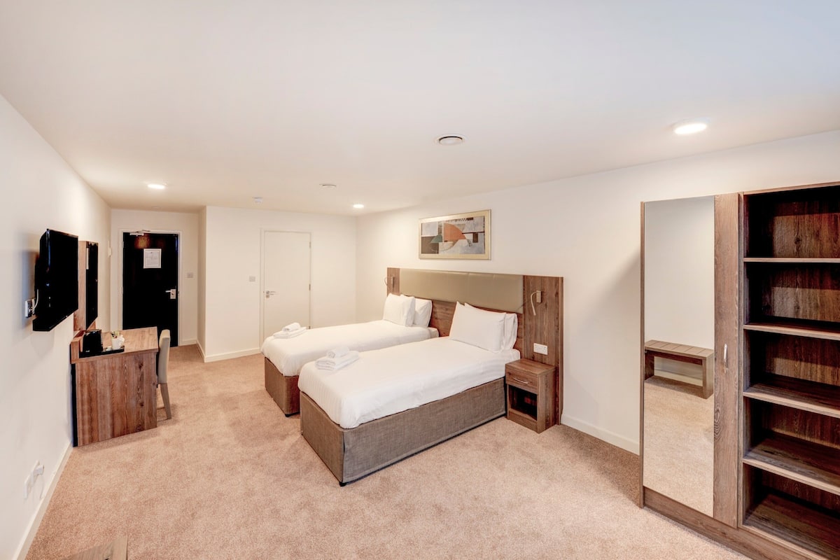 Une chambre d’hôtel avec deux lits, une armoire en bois, un bureau avec une chaise et un téléviseur fixé au mur.