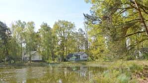 A serene lakeside setting with two houses, one with a green roof and another with a white exterior, surrounded by lush greenery and trees. - Holiday Home in Stramproy Near Nature Reserve (Stramproy)