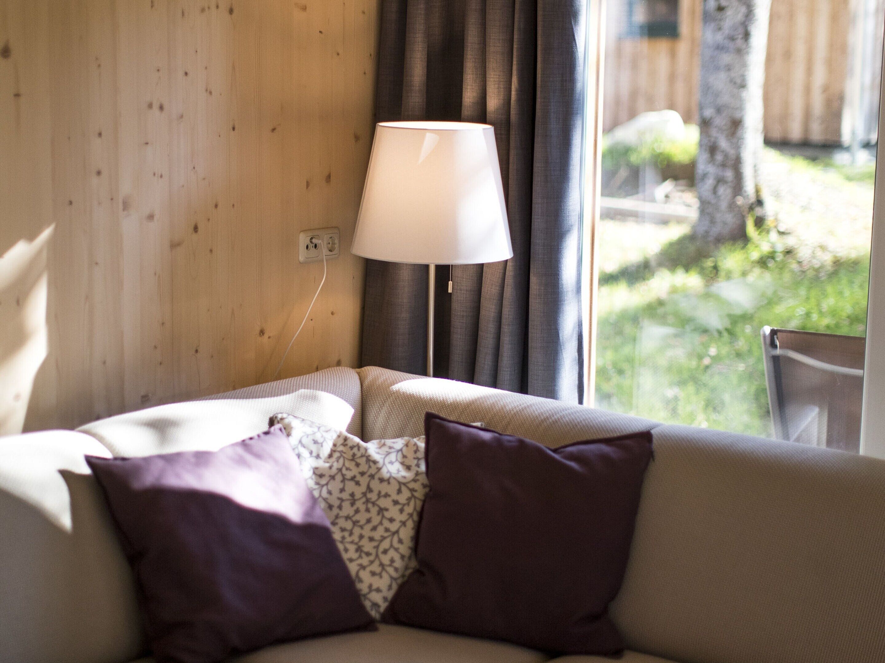 A cozy room with a wooden wall, a window with curtains, a sofa with patterned cushions, and a table lamp.