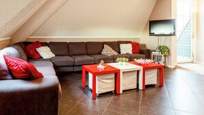 Un salon moderne avec un canapé d’angle marron, des coussins rouges et blancs, une table basse rouge et un téléviseur à écran plat fixé au mur.