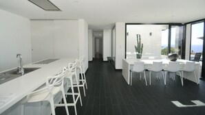 Une cuisine moderne avec un long comptoir et des chaises blanches, un coin repas avec une table et des chaises, et une vue dégagée grâce à de grandes fenêtres.