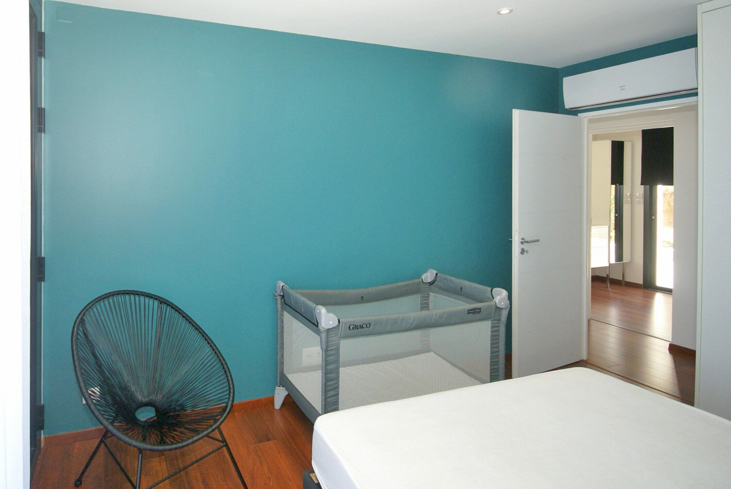 A room with teal walls, a white bed, a Graco playpen, and a black chair.