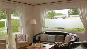 A living room with a sofa, armchair, and a coffee table. There is a window with curtains, a lamp, and a view of a road and greenery outside. - Tidy Chalet with Microwave near Wadden Sea (Tzummarum)