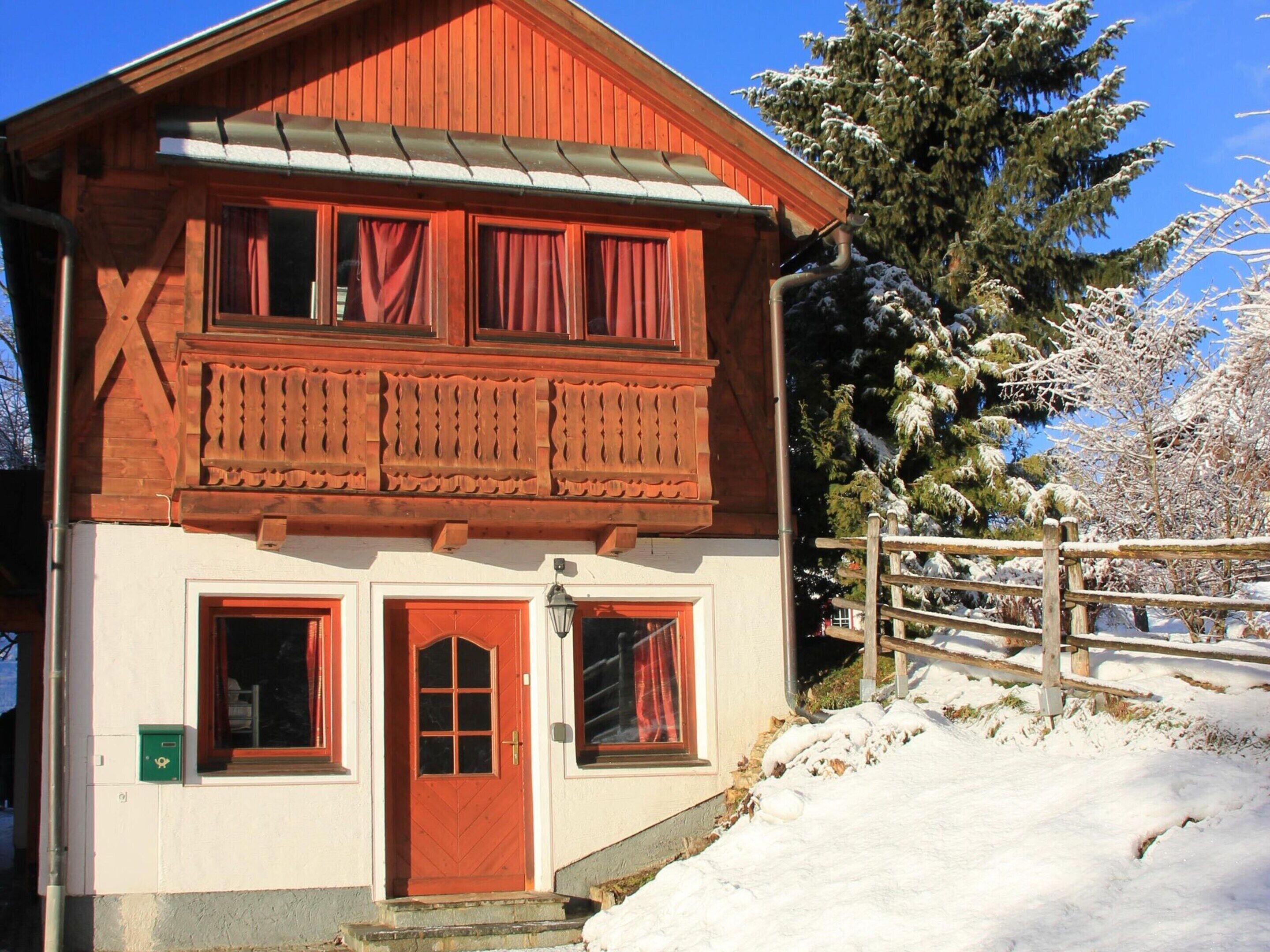 Ein zweistöckiges Holzhaus mit roter Tür und Fenstern, umgeben von Schnee und Bäumen.
