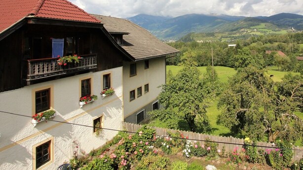 Un chalet traditionnel avec un toit rouge, un balcon en bois orné de jardinières fleuries, et une vue sur une vallée verdoyante et des montagnes.