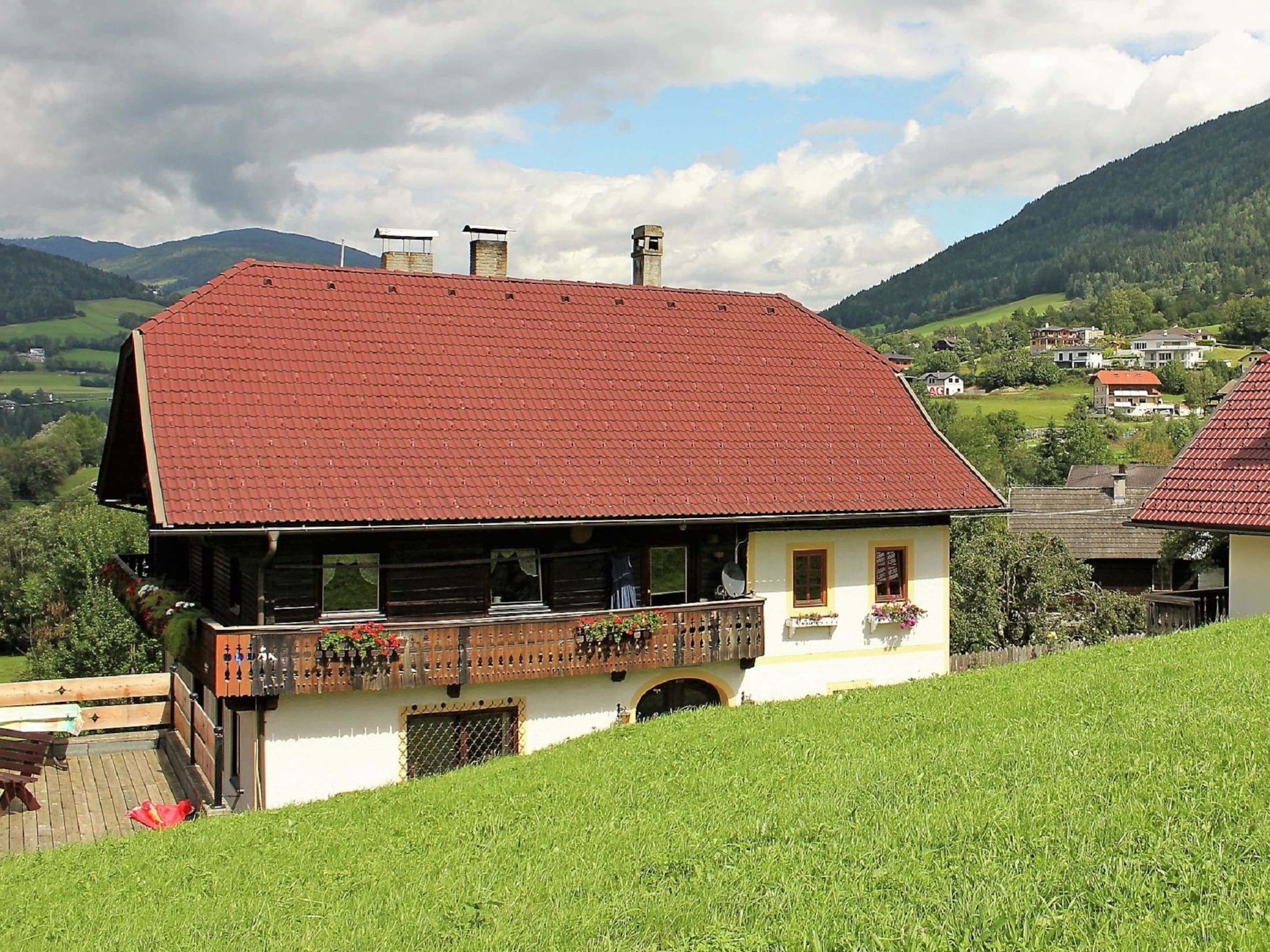 Ein traditionelles Chalet mit geneigtem roten Dach, einem hölzernen Balkon mit Blumenkästen und Blick auf ein grünes Tal und Berge im Hintergrund.