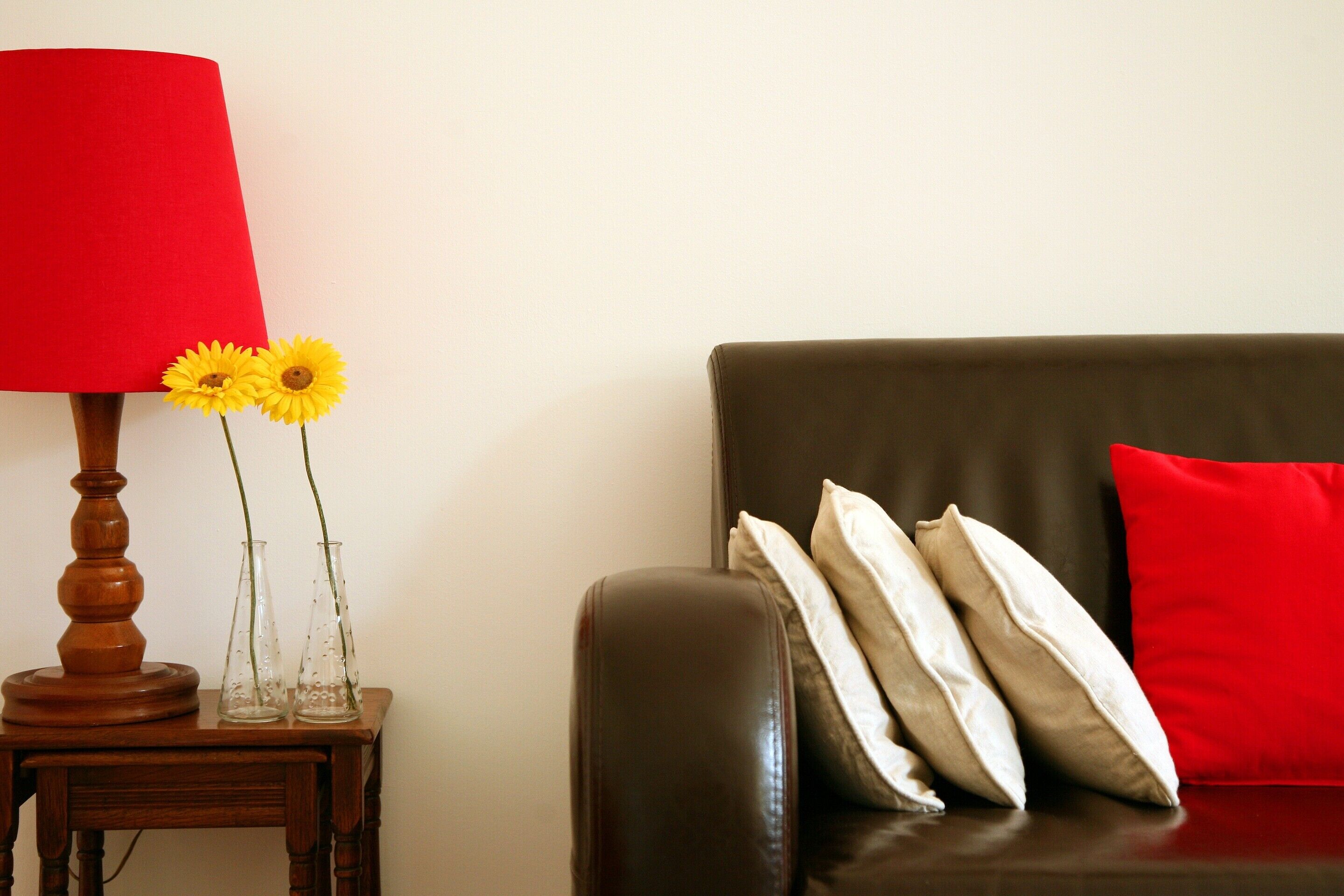 A brown leather sofa with white and red cushions, a wooden side table with a red lampshade and two clear vases with yellow flowers.
