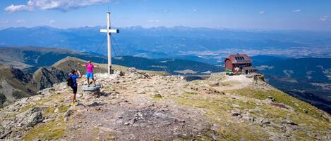 Um pico montanhoso com uma cruz, uma pequena casa com painéis solares e uma vista desobstruída de montanhas ao longe.