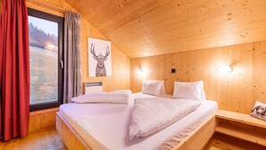 A wooden cabin room with a bed, a window with curtains, a wall-mounted light, and a framed picture. - Chalet in Hohentauern With Sauna (Hohentauern)