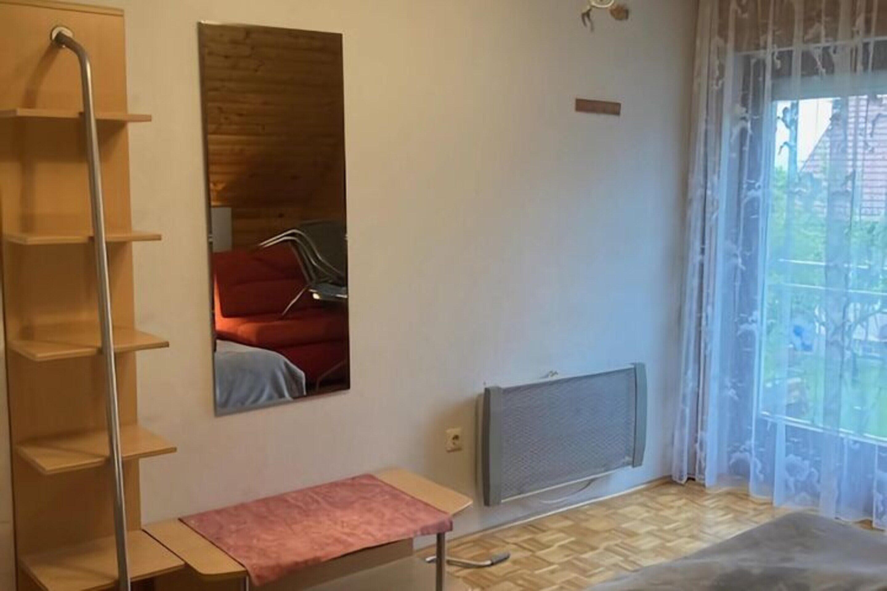 Une pièce avec un plancher en bois, un radiateur, une fenêtre avec des rideaux légers, un miroir qui reflète une chaise rouge et une étagère en métal.