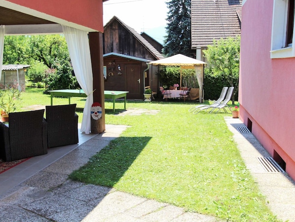 A covered patio area with a table and chairs, a grassy lawn, and a wooden building in the background.
