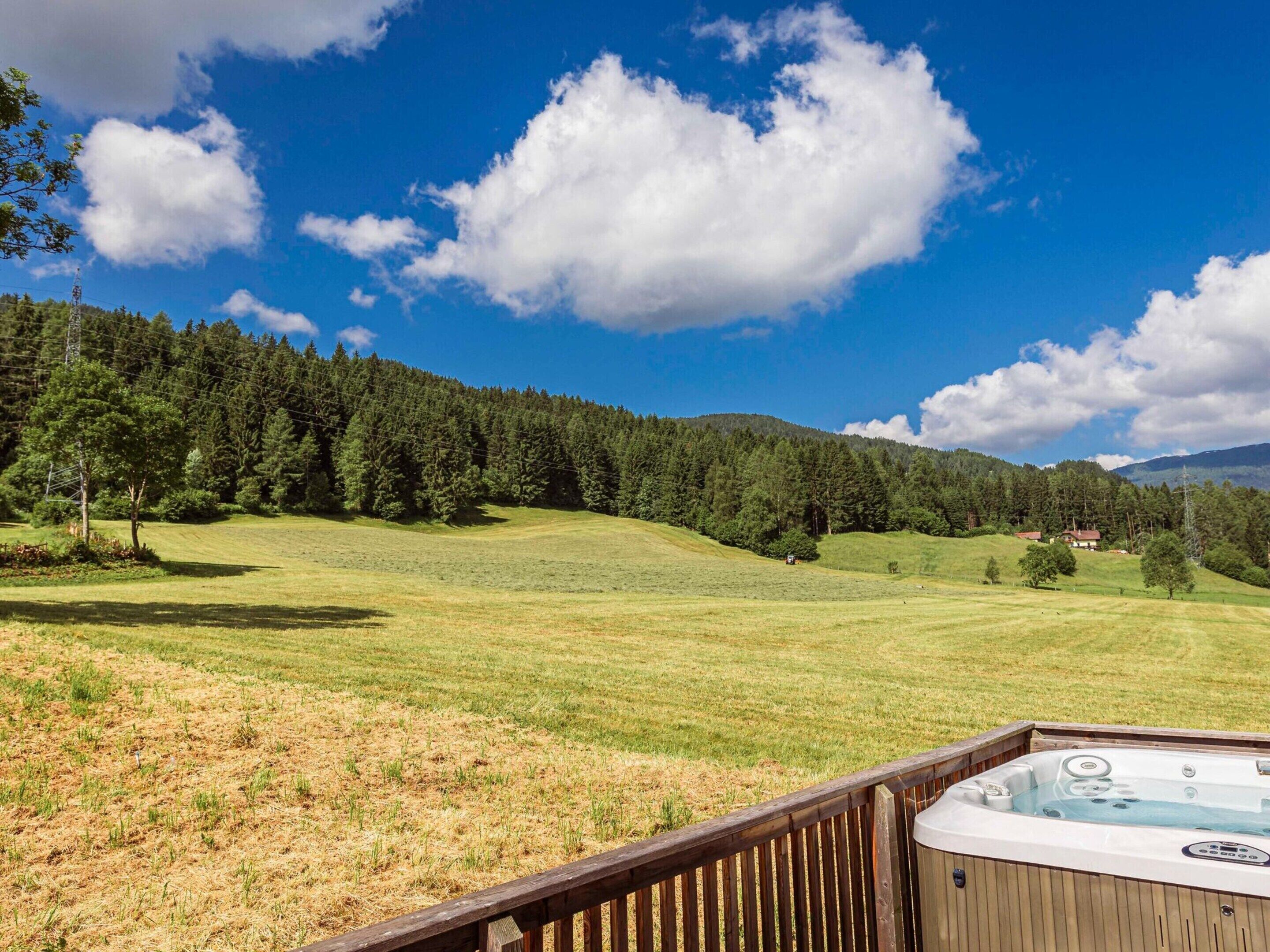 A hot tub on a wooden deck overlooking a grassy field with trees and a house in the distance under a clear blue sky.