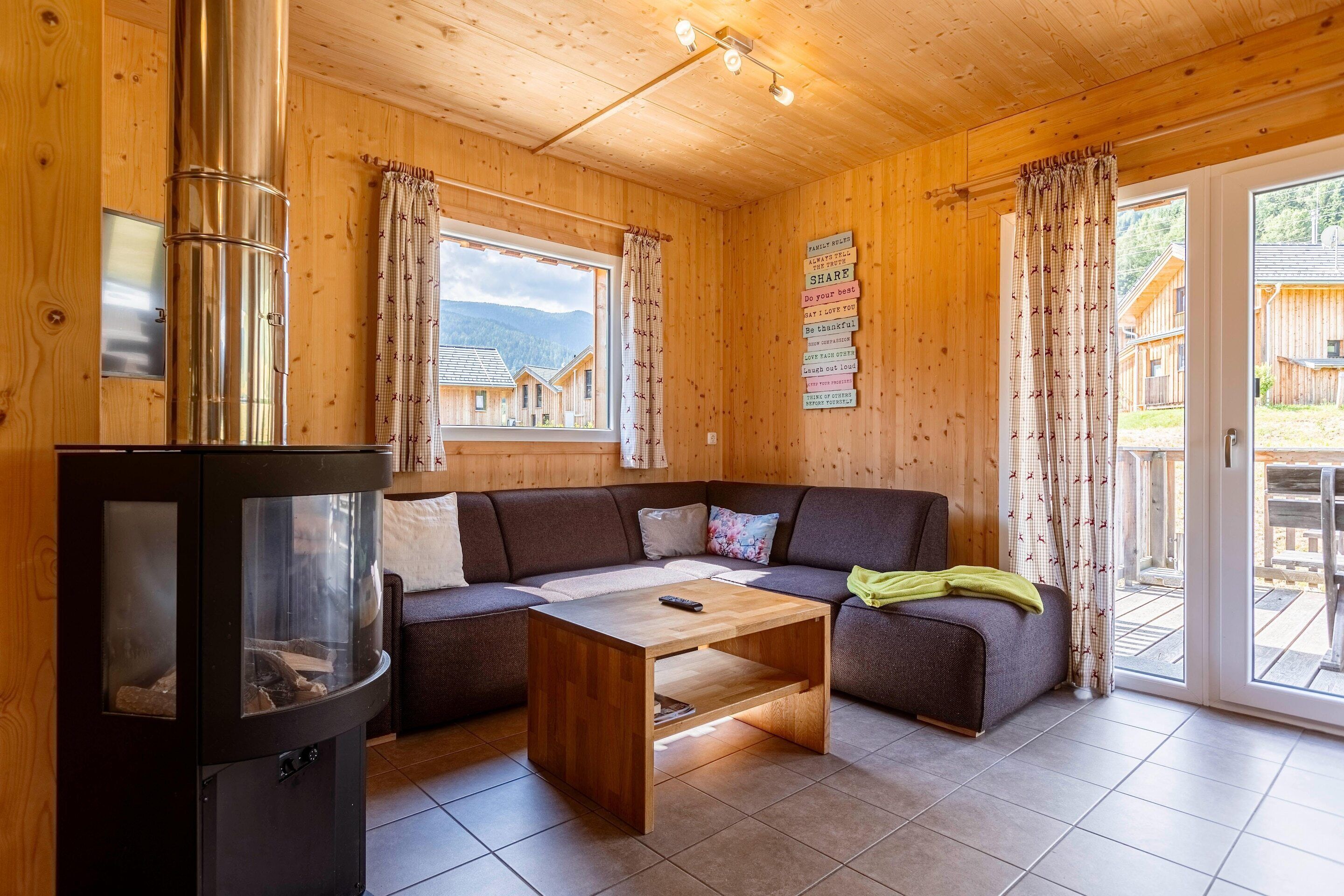 A wooden cabin interior with a sofa, a wooden coffee table, a fireplace, and a view of cabins through the windows.