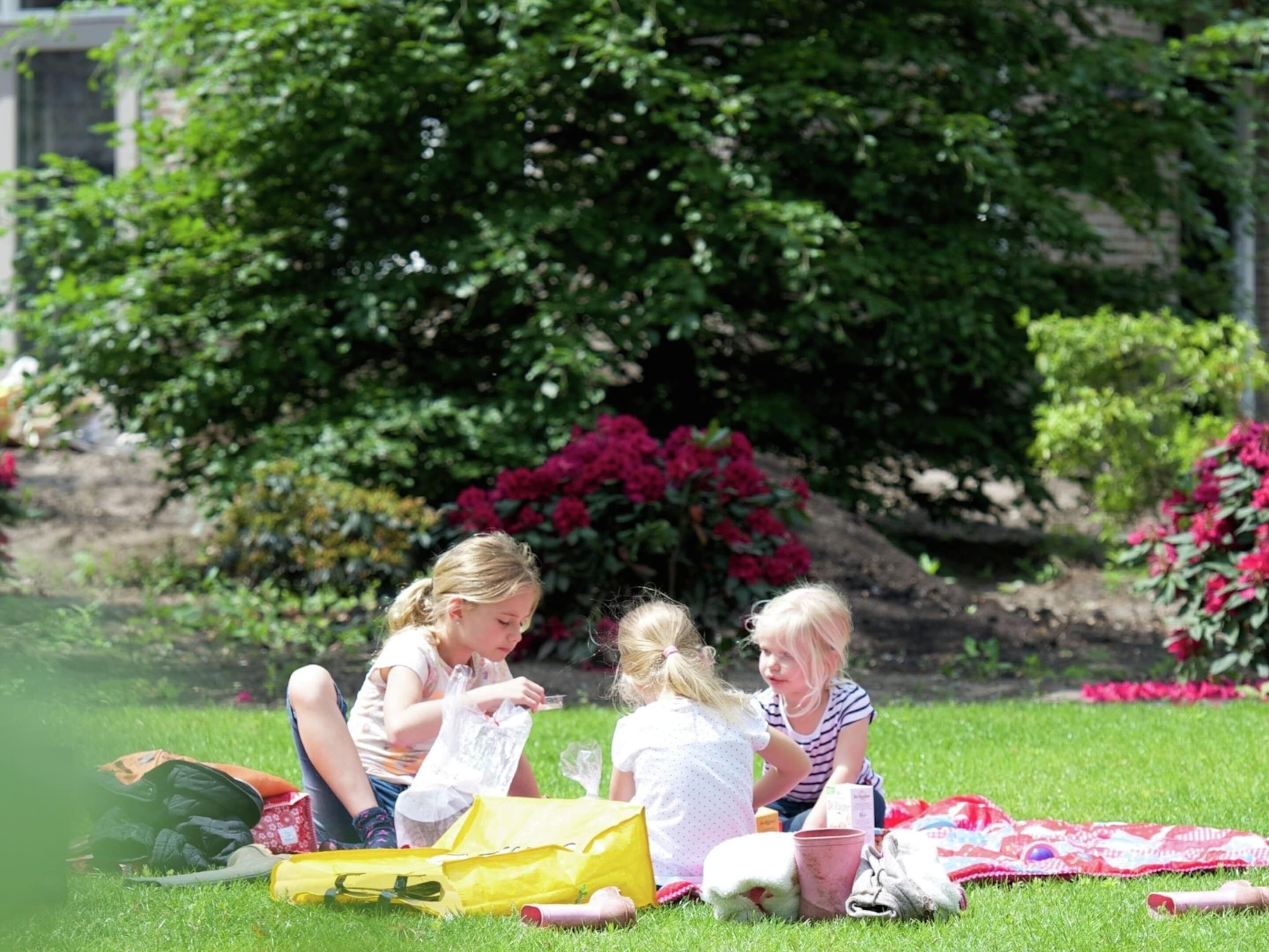 Three children sitting on grass in a park with flowers and trees in the background.