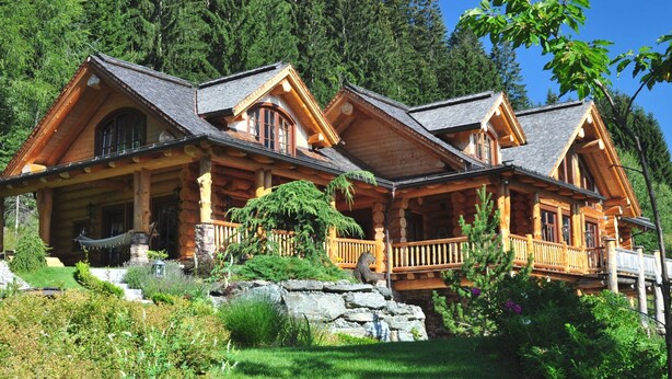 A log cabin with a porch, stone foundation, and a forest backdrop.