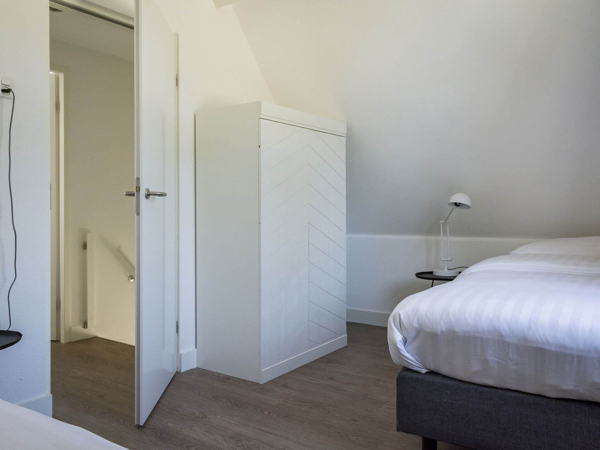 Een slaapkamer met een bed, nachtkastje, lamp, kledingkast en een open deur naar een andere kamer.