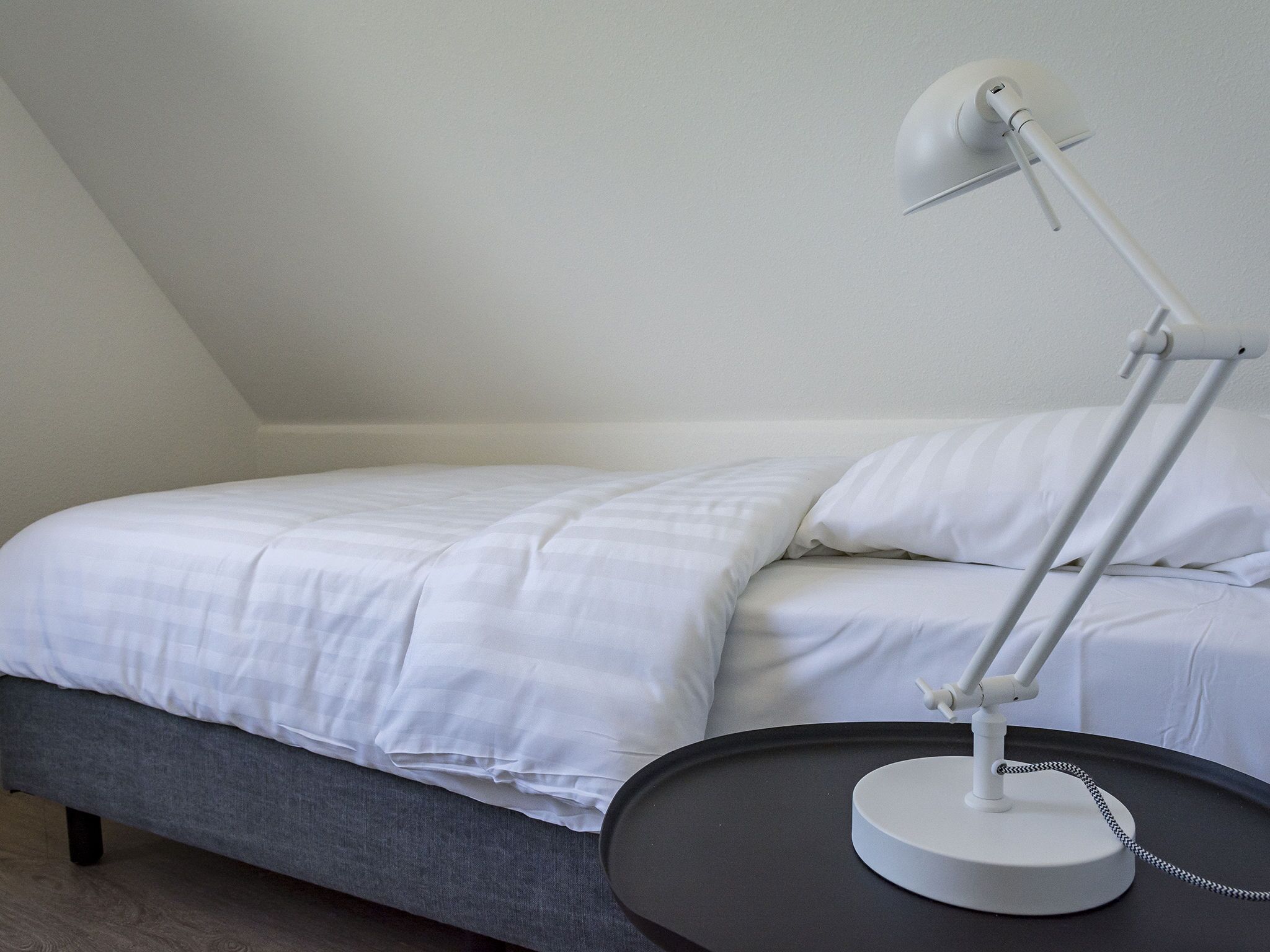 A neatly made bed with white linens, a gray upholstered headboard, and a white desk lamp on a round table beside the bed.